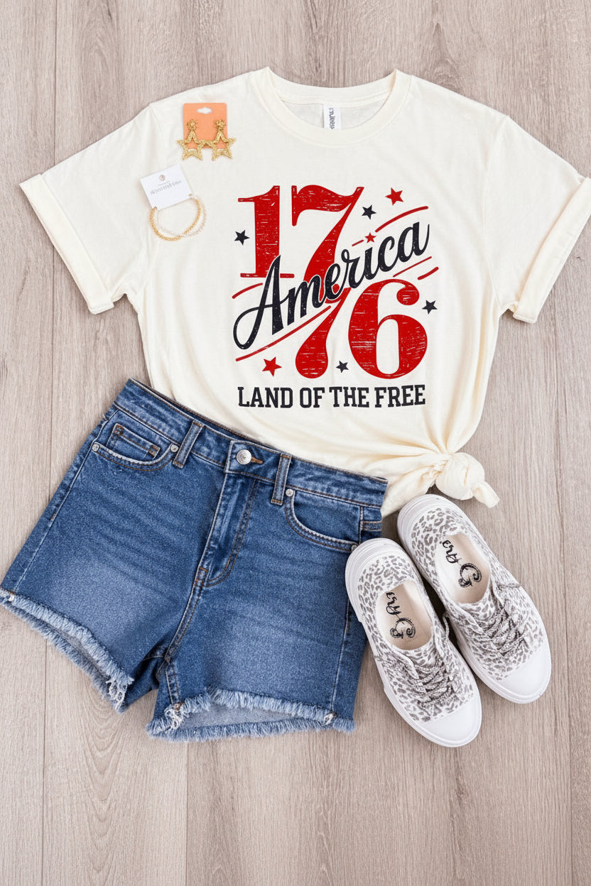 America 1776 patriotic graphic tee on cream Tultex shirt styled with denim shorts and sneakers flat lay