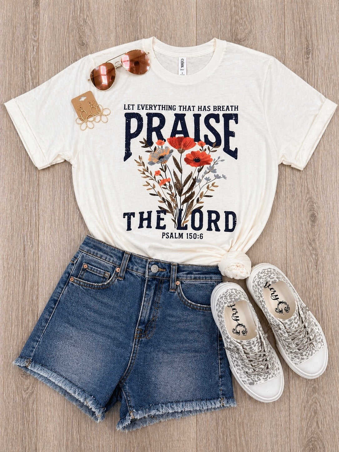 White t-shirt with 'Praise the Lord' design, denim shorts, and white sneakers on a wooden surface.