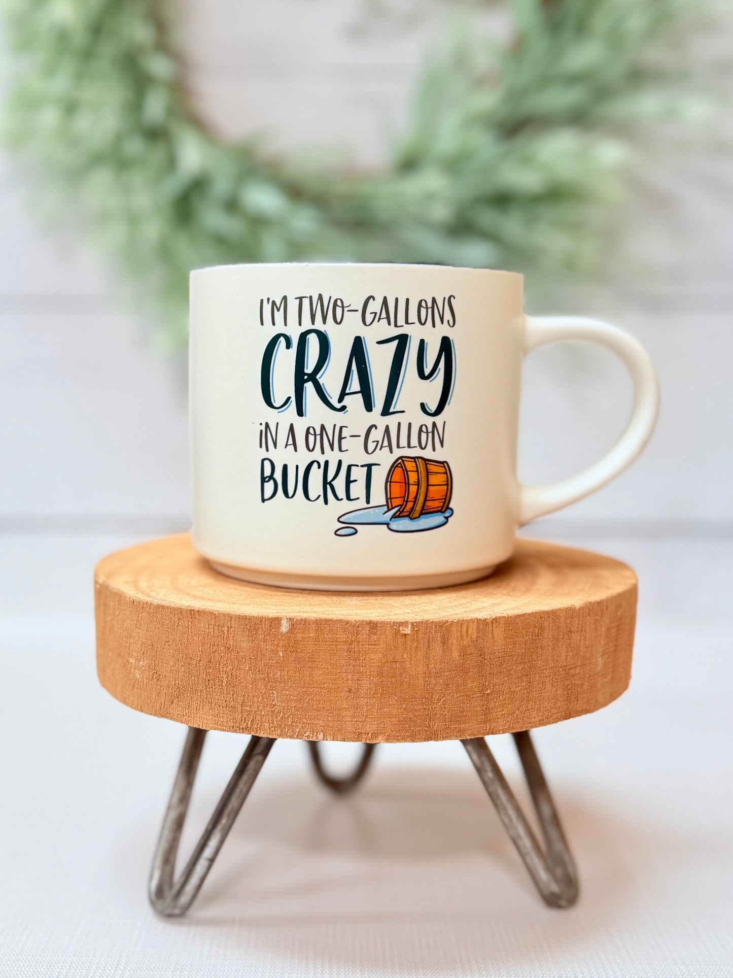 Two Gallons of Crazy coffee mug with humorous text styled on a small wooden stool with greenery background