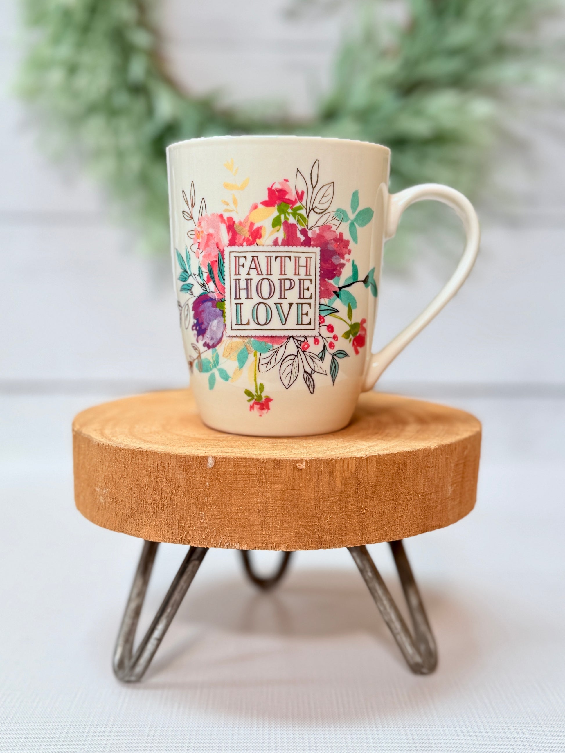 Faith Hope Love coffee mug with colorful floral design styled on a wooden stool with greenery background