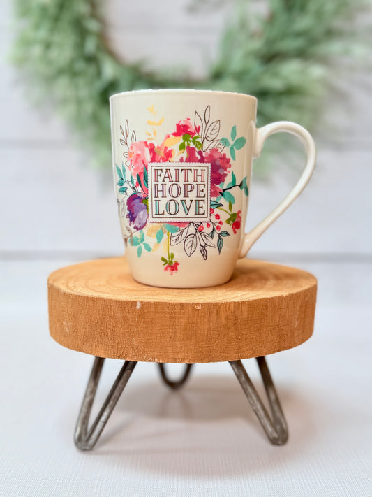 Faith Hope Love coffee mug with colorful floral design styled on a wooden stool with greenery background
