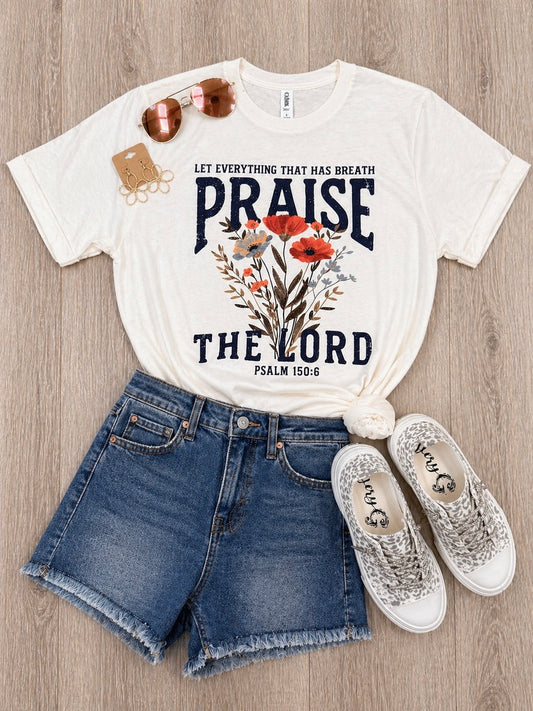 White t-shirt with 'Praise the Lord' design, denim shorts, and white sneakers on a wooden surface.